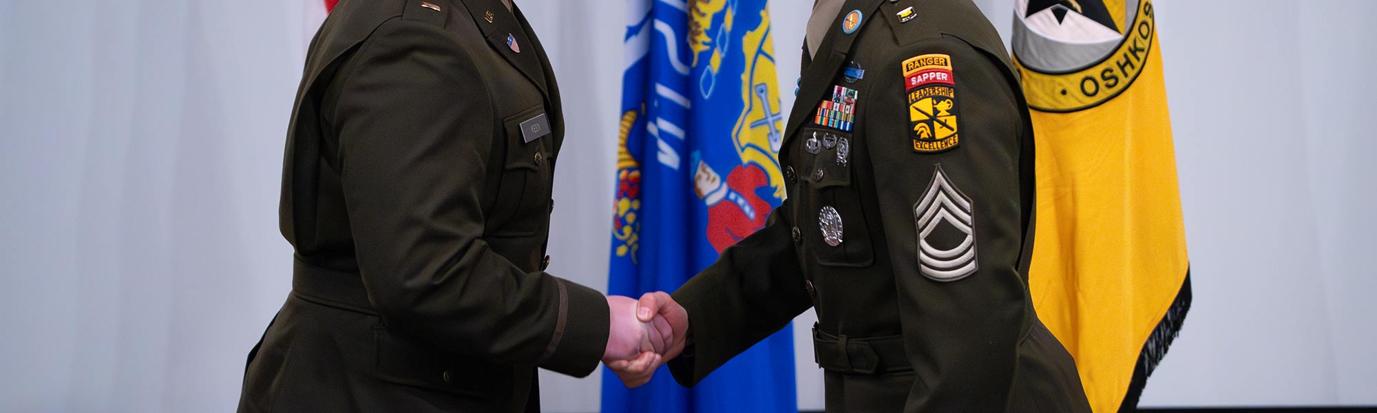 Two military members shake hands in front of flags