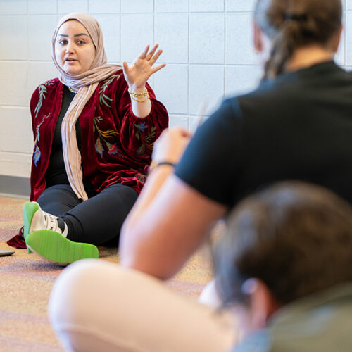 A professor sits with their students on the floor