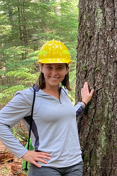 Sarah Anderson stands in a forest in a hard hat resting her hand on a tree.
