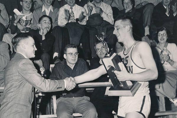 A basketball player shakes hands with a person in a suit as fans cheer.