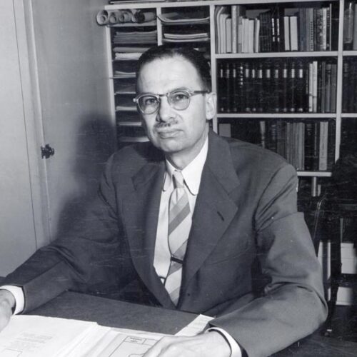 A man in a suit sits at a desk with many books behind him