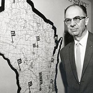 Harold stands in front of a map of Wisconsin with various flags on station points.