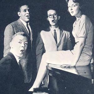 A group of singers gather to sing near a piano