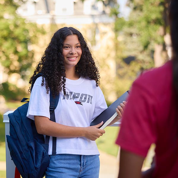 A student in white shirt with backpack holds books as she smiles at a friend